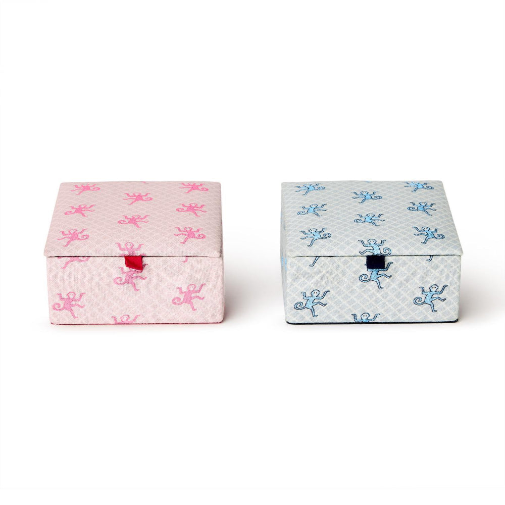 Two small square decorative boxes on a white background, one pink with a repeating pink motif and a red tab closure, and one light blue with a repeating blue motif and a dark blue tab closure.