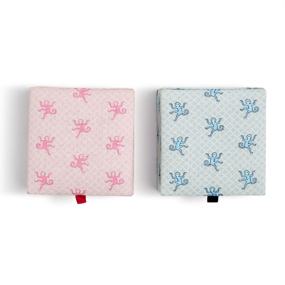 Two square baby footmuffs side by side on a white background. The left one is light pink with a repeating pink cherub pattern and a small red tab.