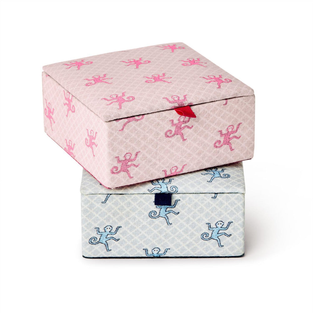 Two stacked fabric-covered storage boxes on a white background. The top box is light pink with a repeating pink monkey print and a small red ribbon clasp.