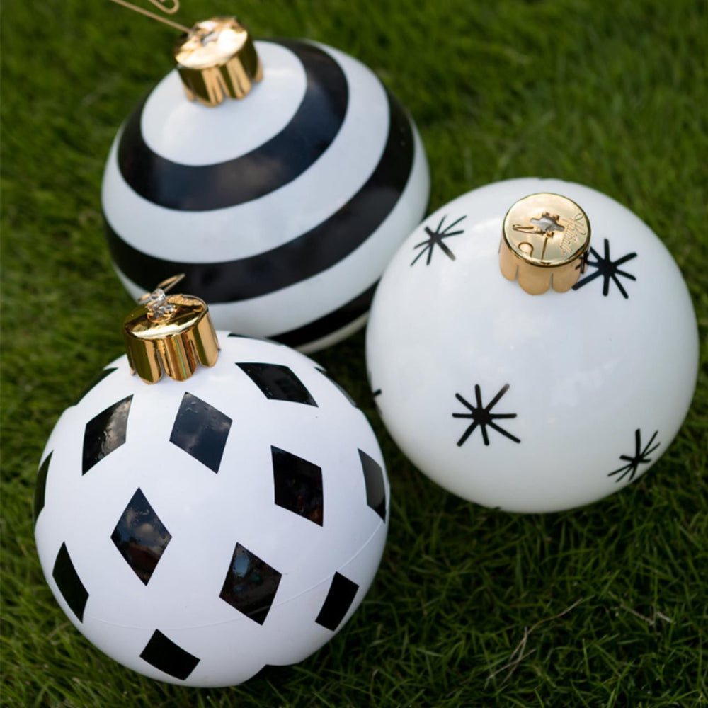Three decorative Christmas ornaments with black and white patterns resting on green grass. One ornament features bold black horizontal stripes, another has a black diamond lattice pattern on a white base, and the third is white with black starburst motifs.
