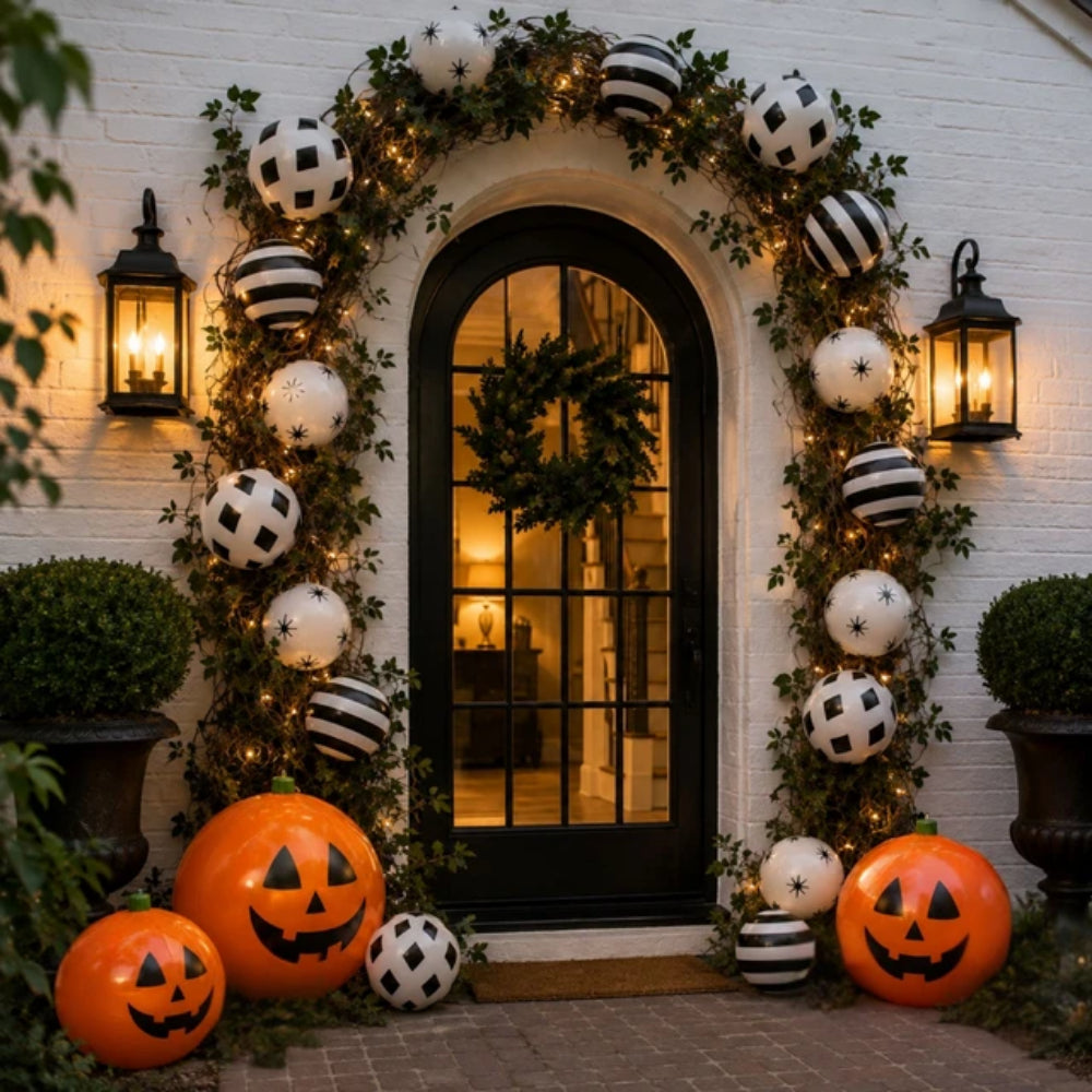 A beautifully decorated front door for Halloween, featuring a black arched door framed by a lush green garland with warm white string lights and black-and-white striped spherical ornaments. Large orange jack-o'-lantern pumpkins are arranged on the stone ground at the base of the door, flanked by potted green shrubs and lit black wall lanterns.