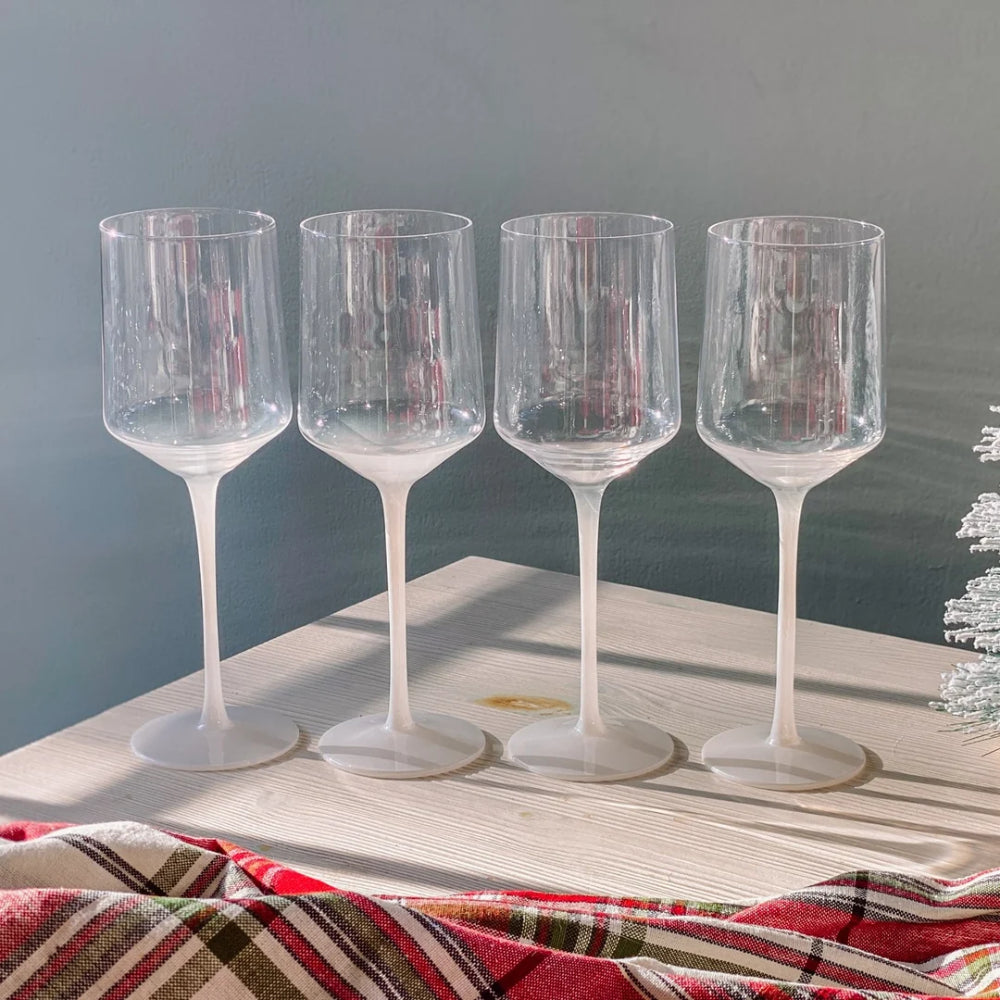 Four clear stemmed wine glasses arranged in a row on a light wooden surface, with a red plaid fabric draped in the foreground and a soft gray wall behind.