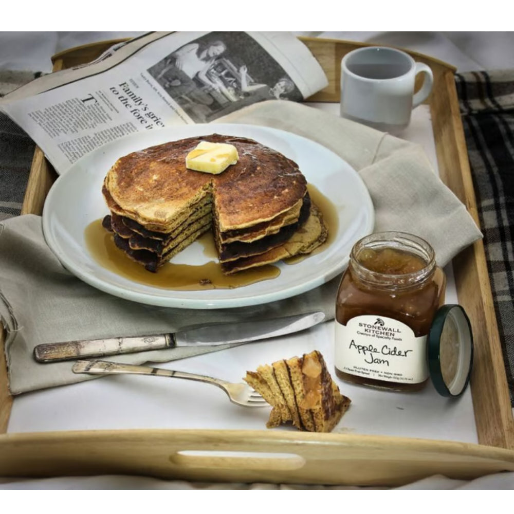A styled breakfast scene on a wooden tray with a plate of pancakes topped with butter and syrup, a jar of Apple Cider Jam, a mug, a knife, a fork, and an open newspaper.