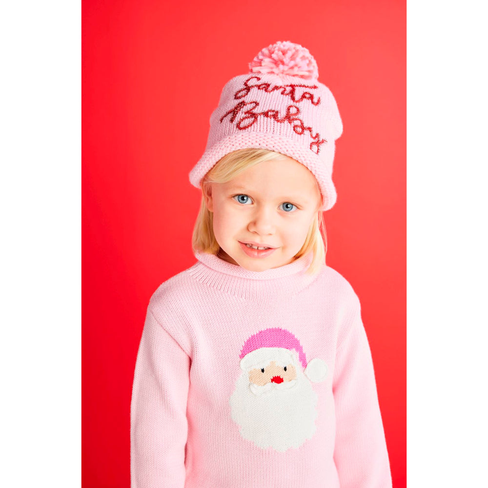 A little girl in a pink sweater with Santa Claus design and a cap with santa baby on a red & white background.