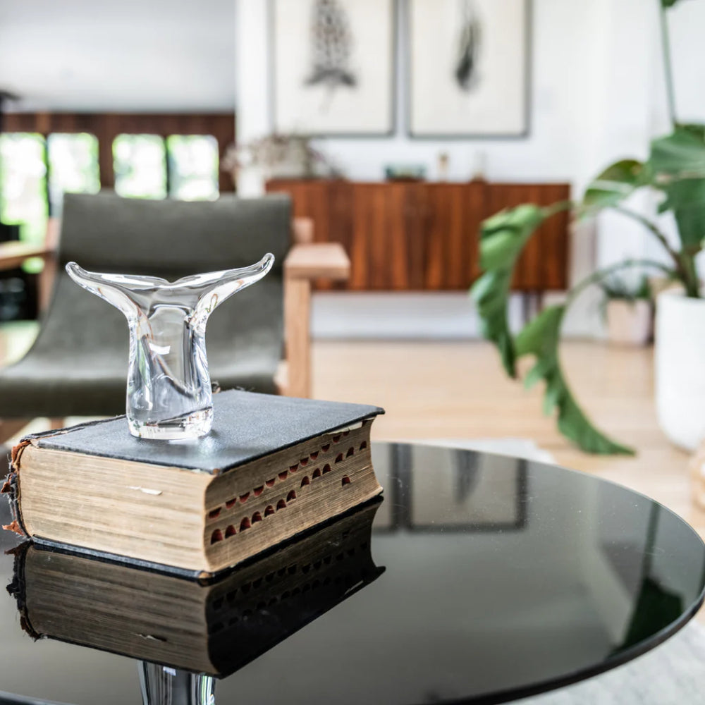 Clear glass Simon Pearce Whale Tail on top of a book on a black coffee table in a room with funiture and plants in it with with painted walls and artwork on it. 