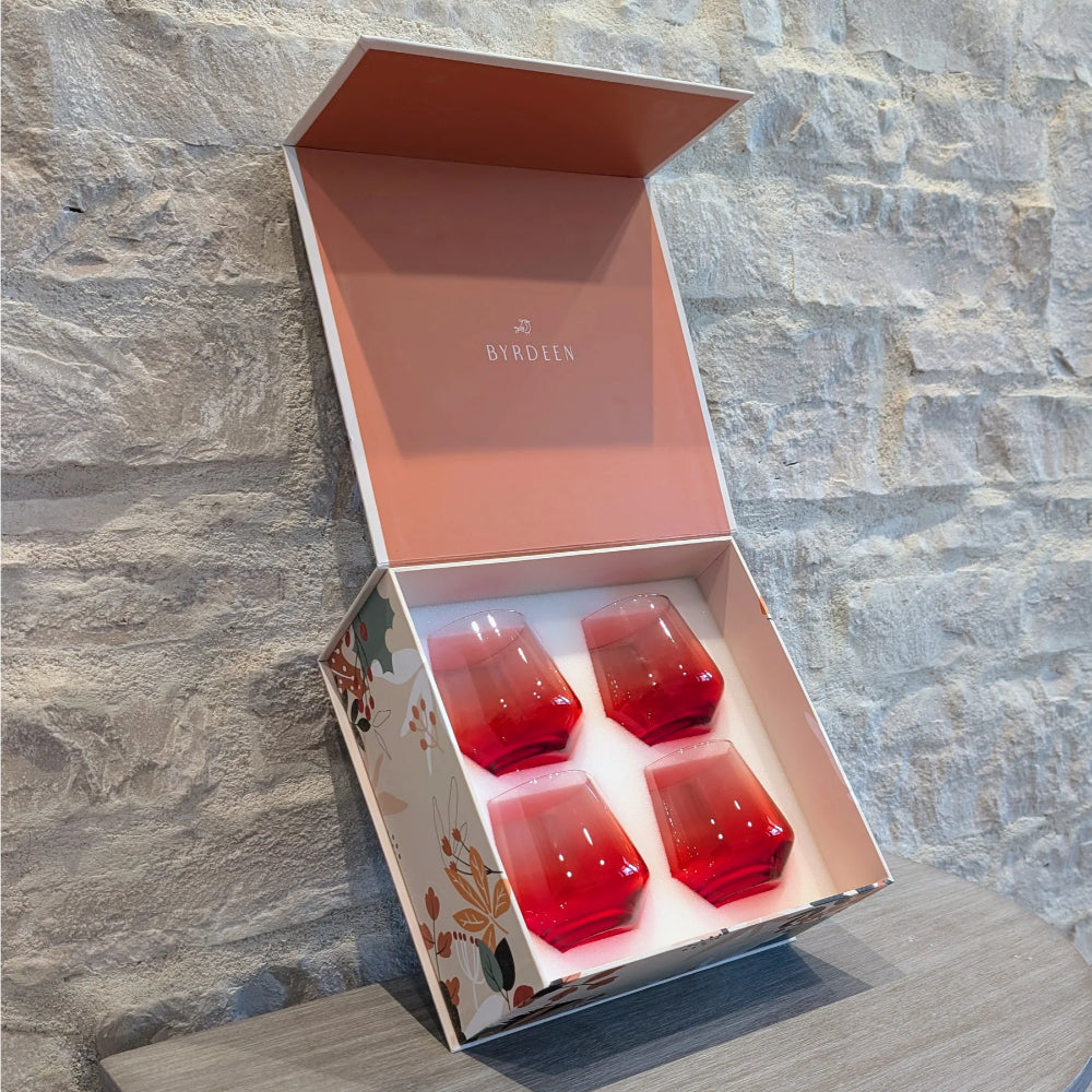 An open decorative gift box containing four small red glass tumblers, set against a light stone wall background. The box has floral detailing on the exterior and the brand name 'BYRDLEN' printed inside the lid.
