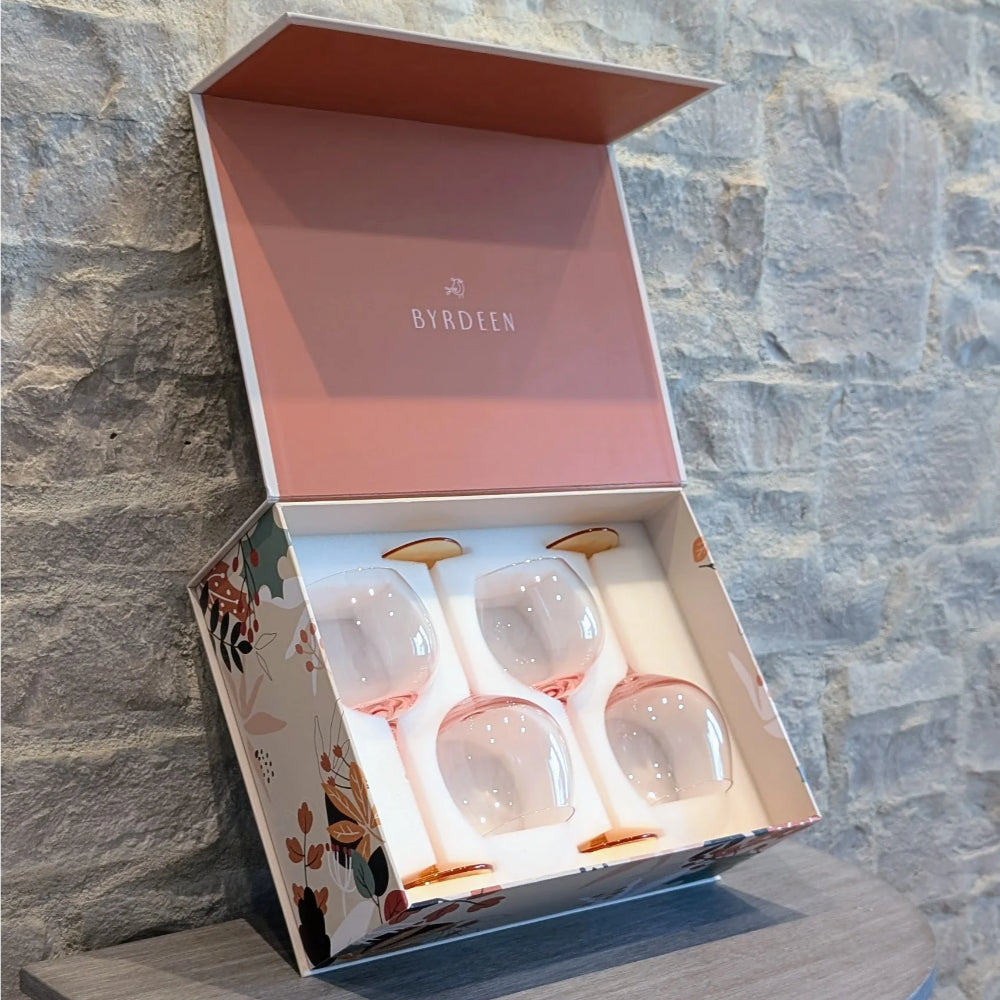 A boxed set of four Byrdeen wine glasses with pale pink stems and gold-toned bases, presented in an open floral gift box against a stone wall background.