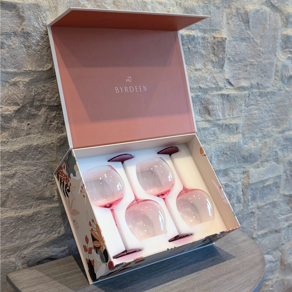 A boxed set of four Byrdeen wine glasses with pale pink stems and pink-tinted bowls, presented in an open floral gift box against a stone wall background.
