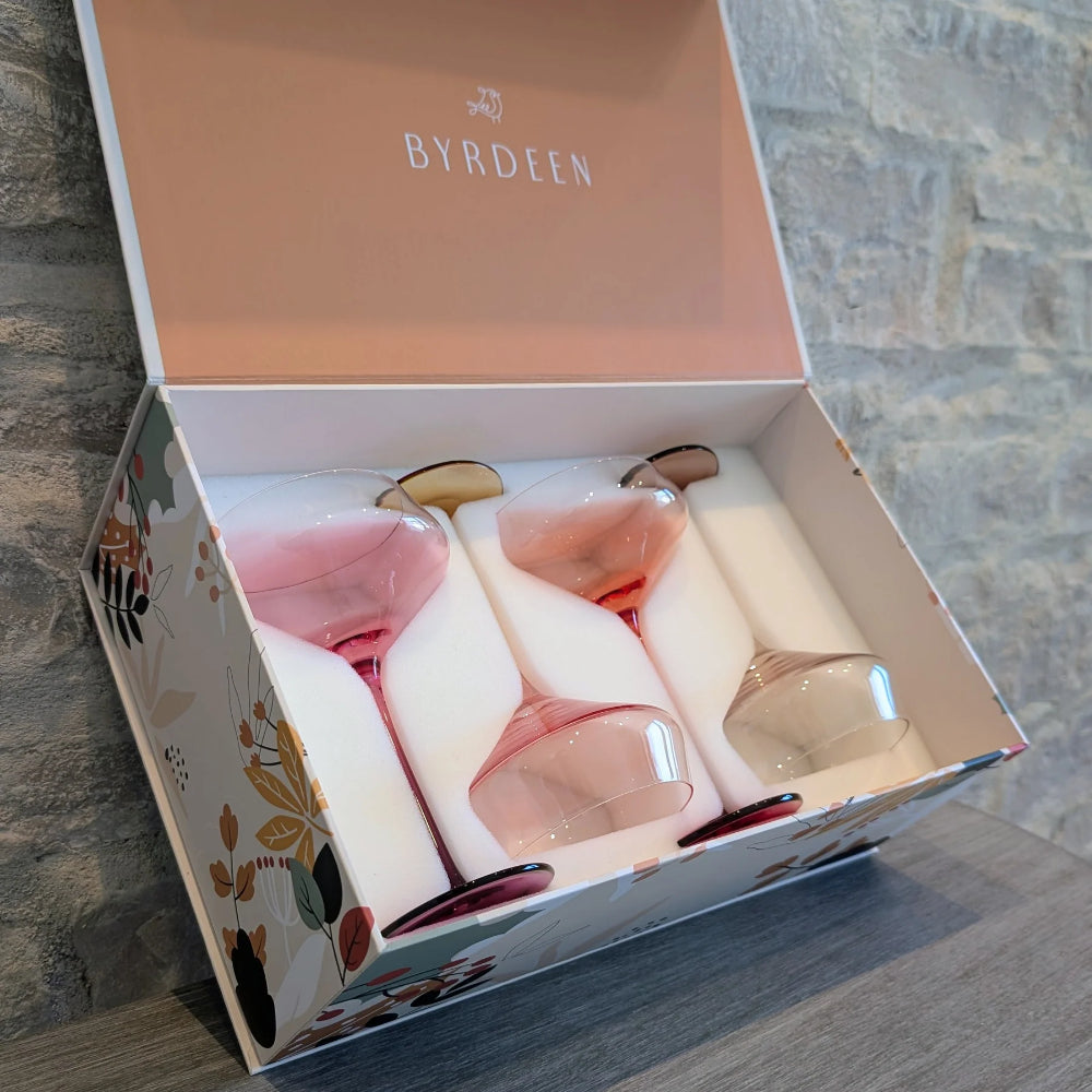 A boxed set of four Byrdeen wine glasses with pale pink tinted glass, presented in a floral-patterned gift box with a peach interior lid, photographed on a tabletop against a stone wall.