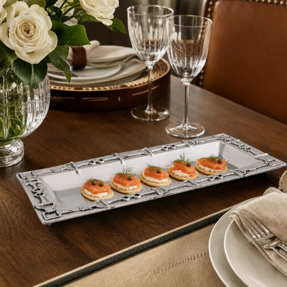 A long rectangular silver serving platter with decorative corner details, holding four small appetizers topped with salmon, set on a dark wood dining table with wine glasses, plates, and a flower vase in the background.