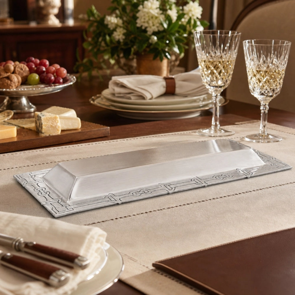 A rectangular silver serving platter with decorative embossed corners, displayed on a beige table runner on a dining table setting with cheese, grapes, plates, and wine glasses.