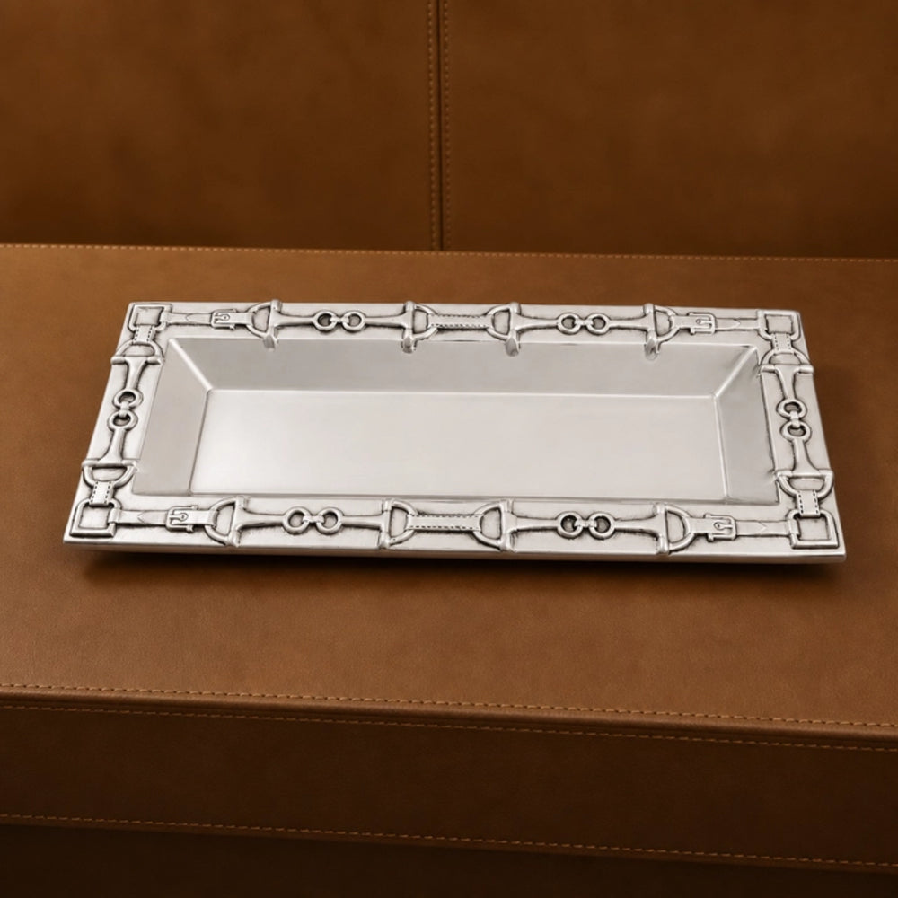A rectangular silver tray with an ornate decorative border featuring horsebit motifs, displayed on a brown leather surface.