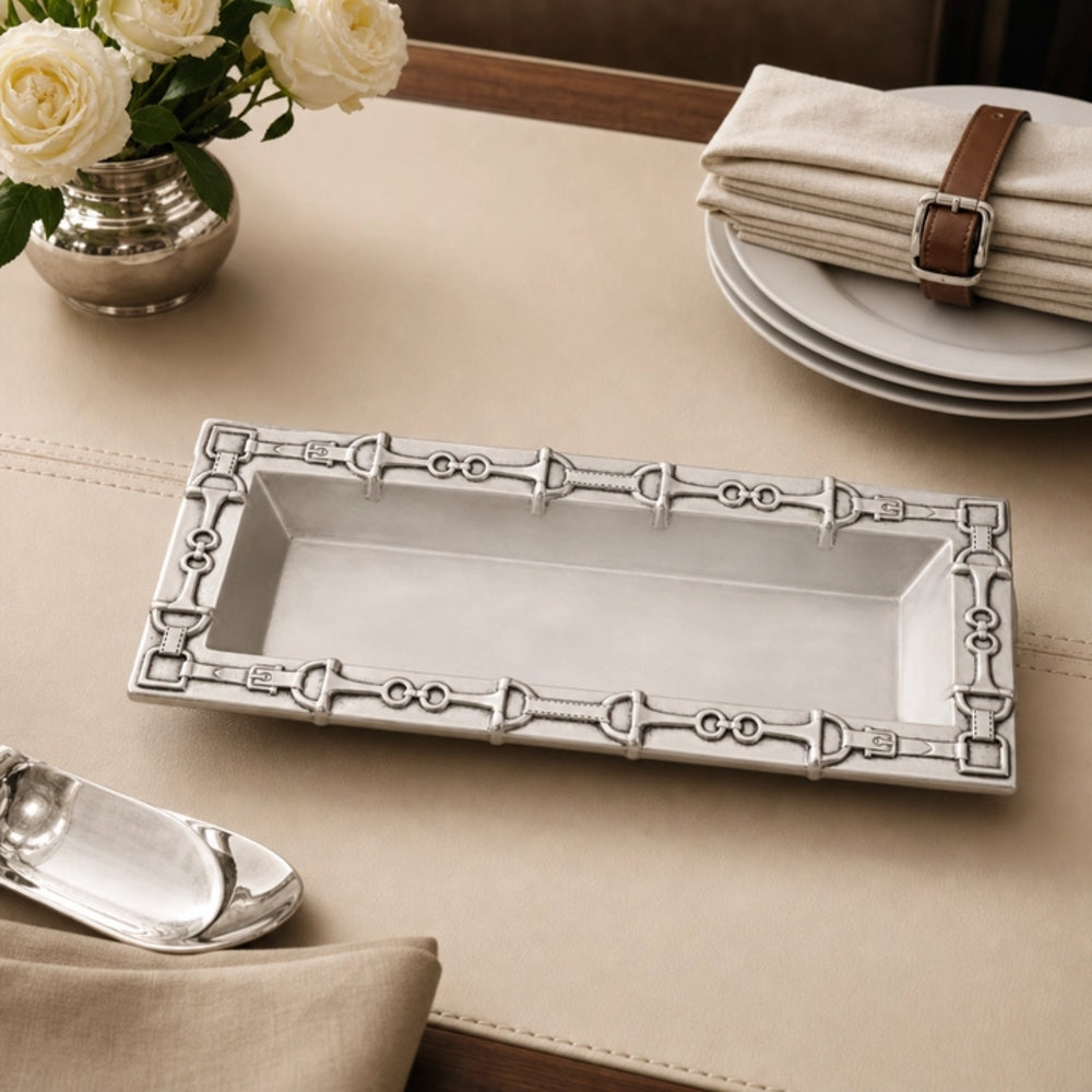 A rectangular silver tray with an ornate decorative border featuring horsebit motifs, styled on a beige table runner with white plates, folded napkins, and white roses in the background.