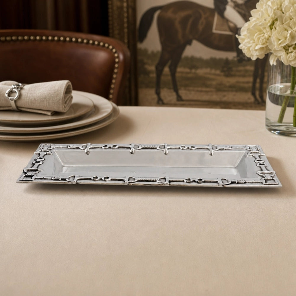A rectangular silver platter with an ornate decorative border, displayed on a beige tablecloth in a dining setting with plates, a chair, and a vase of white flowers.