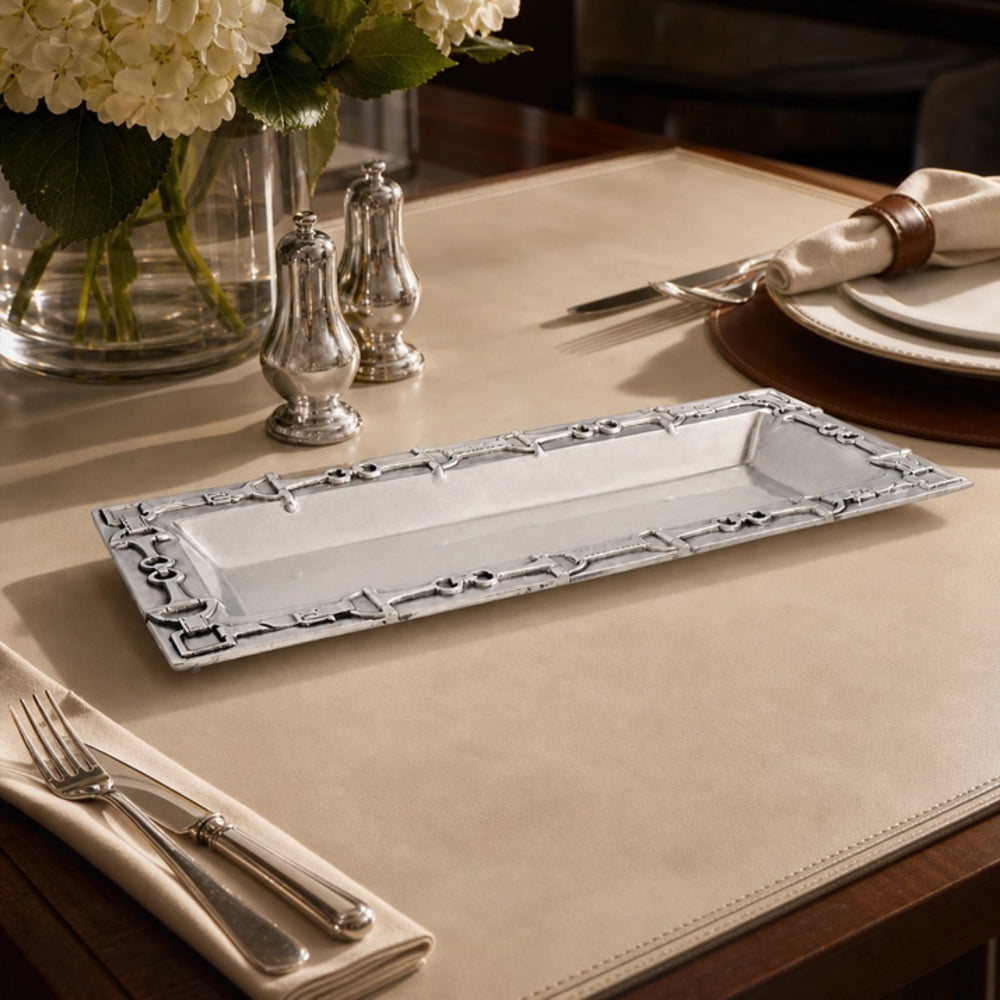 A rectangular silver platter with an ornate raised border, displayed on a beige placemat on a dining table with silverware, salt and pepper shakers, and a flower vase in the background.