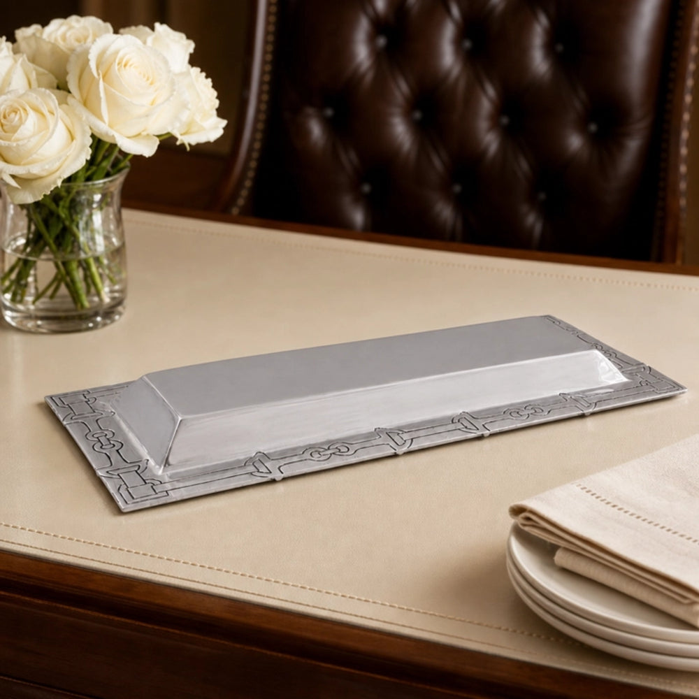 A rectangular silver serving platter with decorative embossed borders, displayed on a beige tablecloth next to a glass vase of white roses and stacked plates.