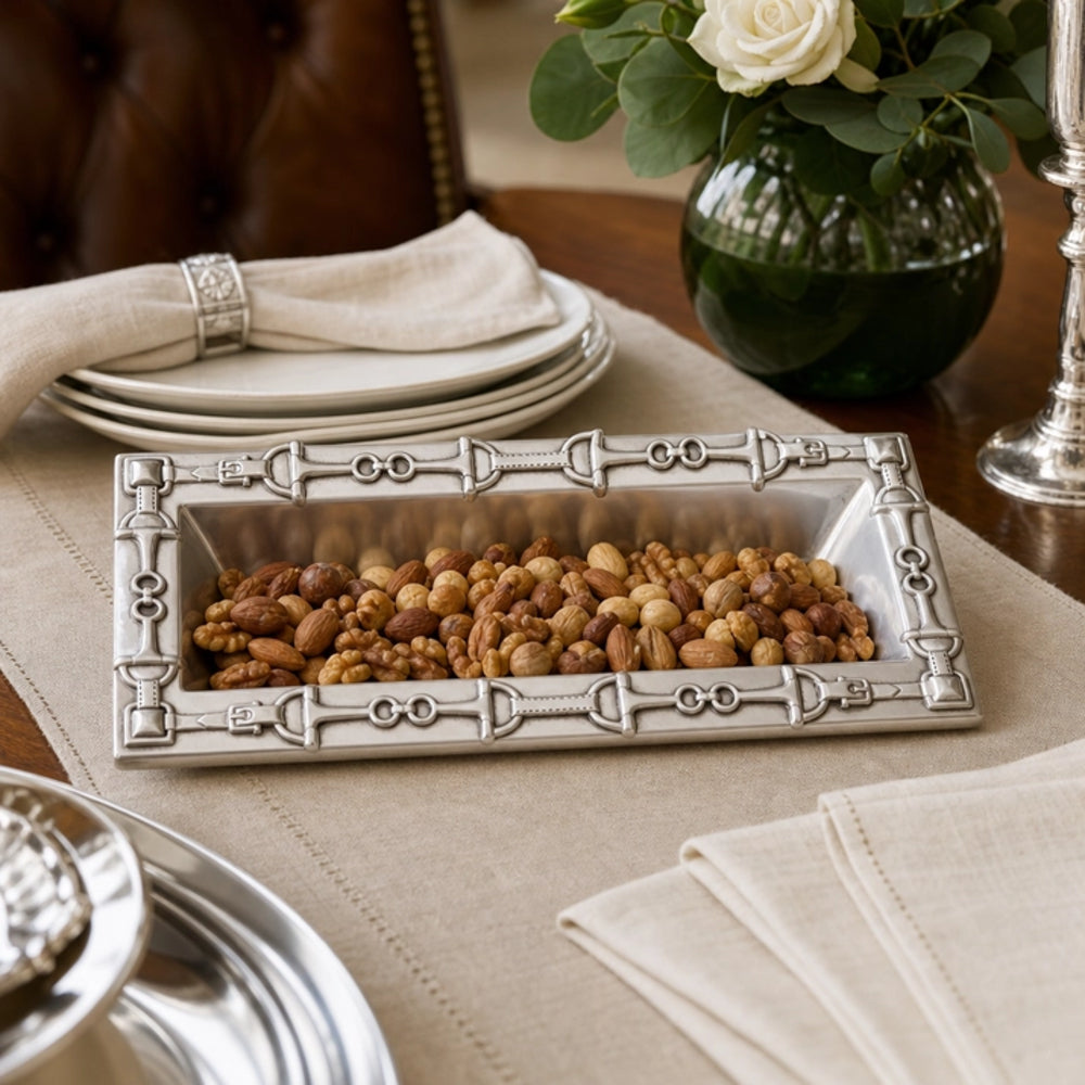 A rectangular silver tray with ornate decorative borders, filled with mixed nuts and dried fruit, styled on a beige tablecloth with plates, folded napkins, and a glass vase of white roses in the background.