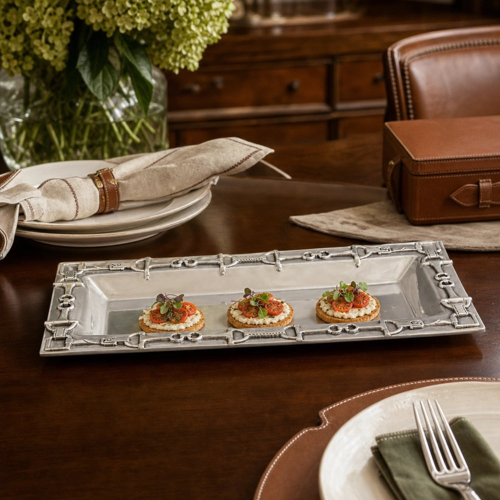 A rectangular silver serving platter with ornate decorative borders, holding three small appetizers on a dark wooden dining table. The scene includes plates, folded napkins, a leather box, and a vase of flowers in a styled interior setting.