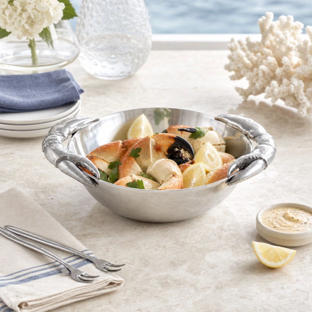 A stainless steel lobster claw serving bowl filled with seafood pasta, set on a light stone tabletop with a napkin, forks, a small dish of sauce, and decorative coral in the background.