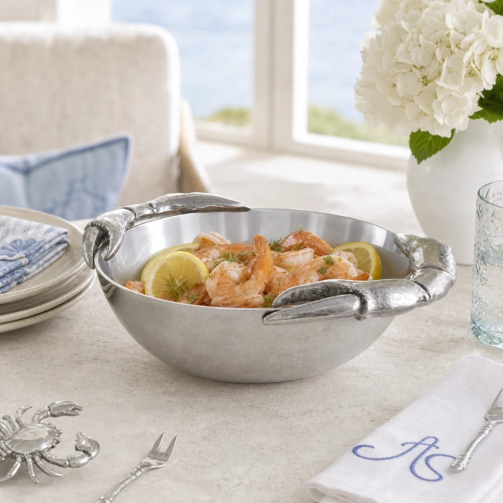A silver metal shrimp bowl with lobster claw handles, filled with cooked shrimp, lemon slices, and herbs, set on a dining table in a bright, coastal-themed room.