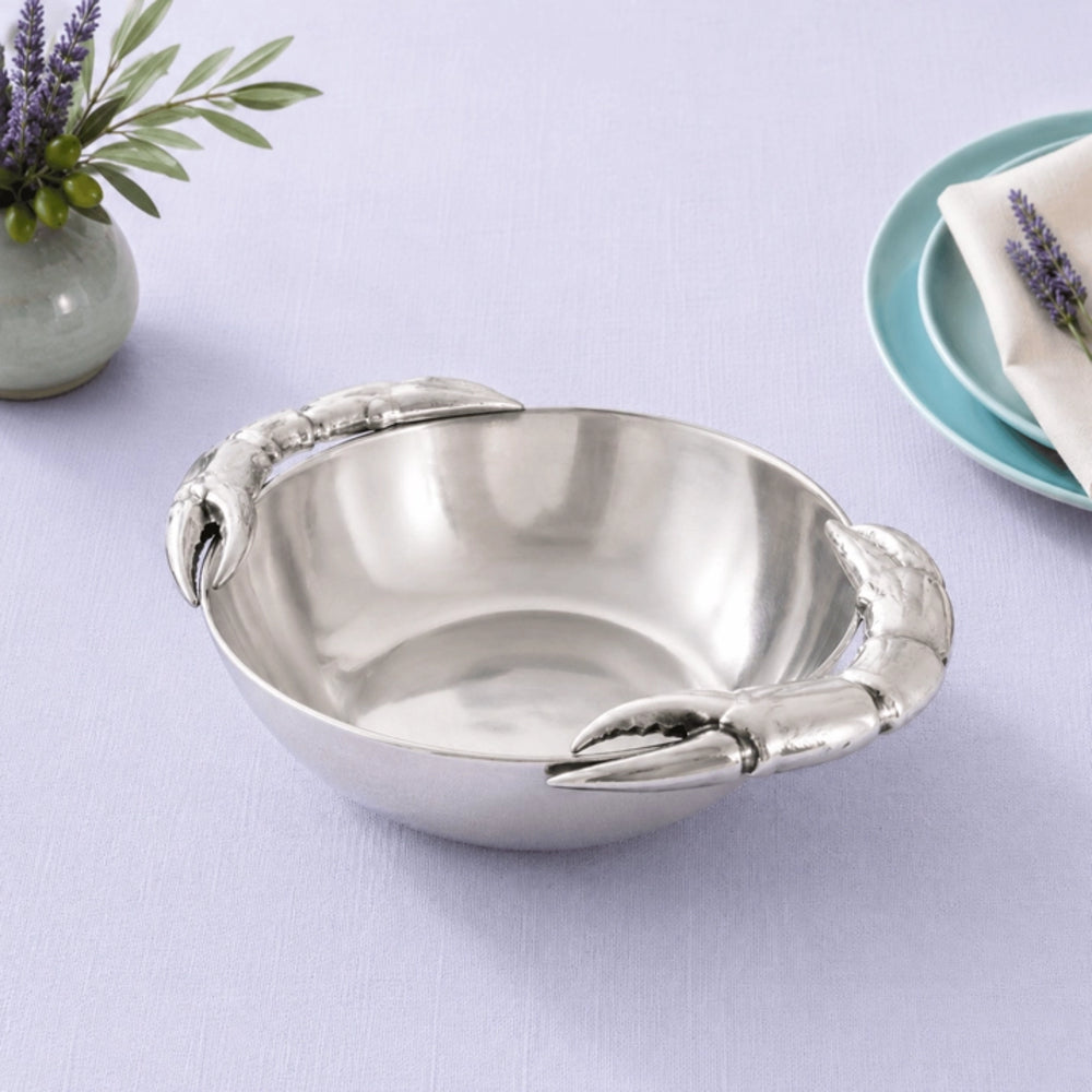 A stainless steel bowl with sculpted lobster claw handles, shown on a light lavender tablecloth with small decorative props including a vase of greenery and plates with lavender.