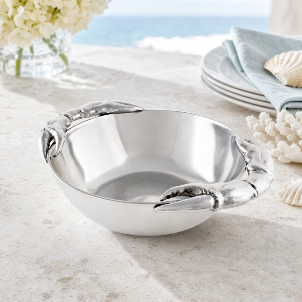 A polished silver bowl with sculpted lobster claw handles, styled on a light stone surface with seashells, plates, and flowers in the background.