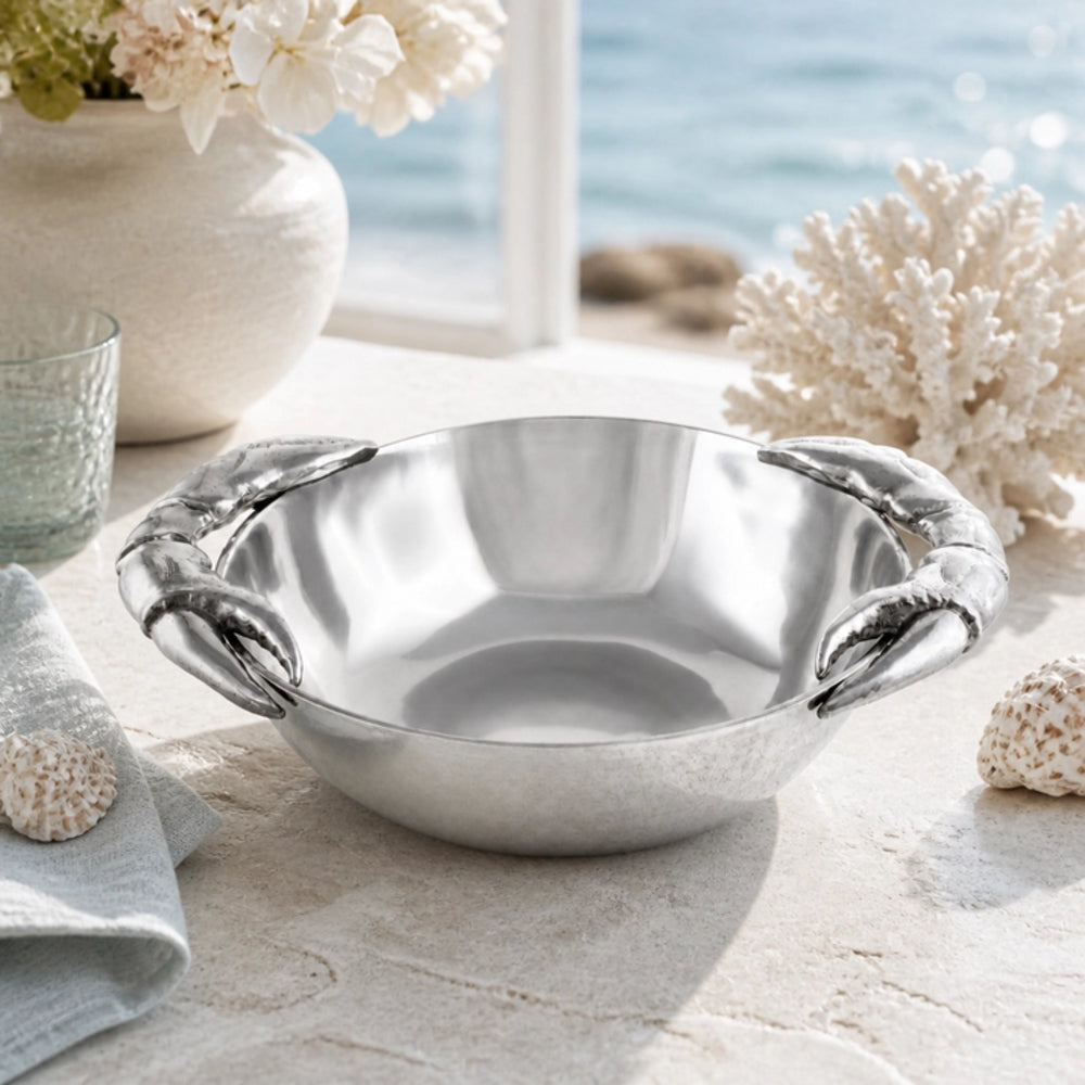 A polished silver metal bowl with sculpted lobster claw handles, styled on a beach-themed surface with seashells, coral, a vase of flowers, and a glass, with a blurred ocean background.