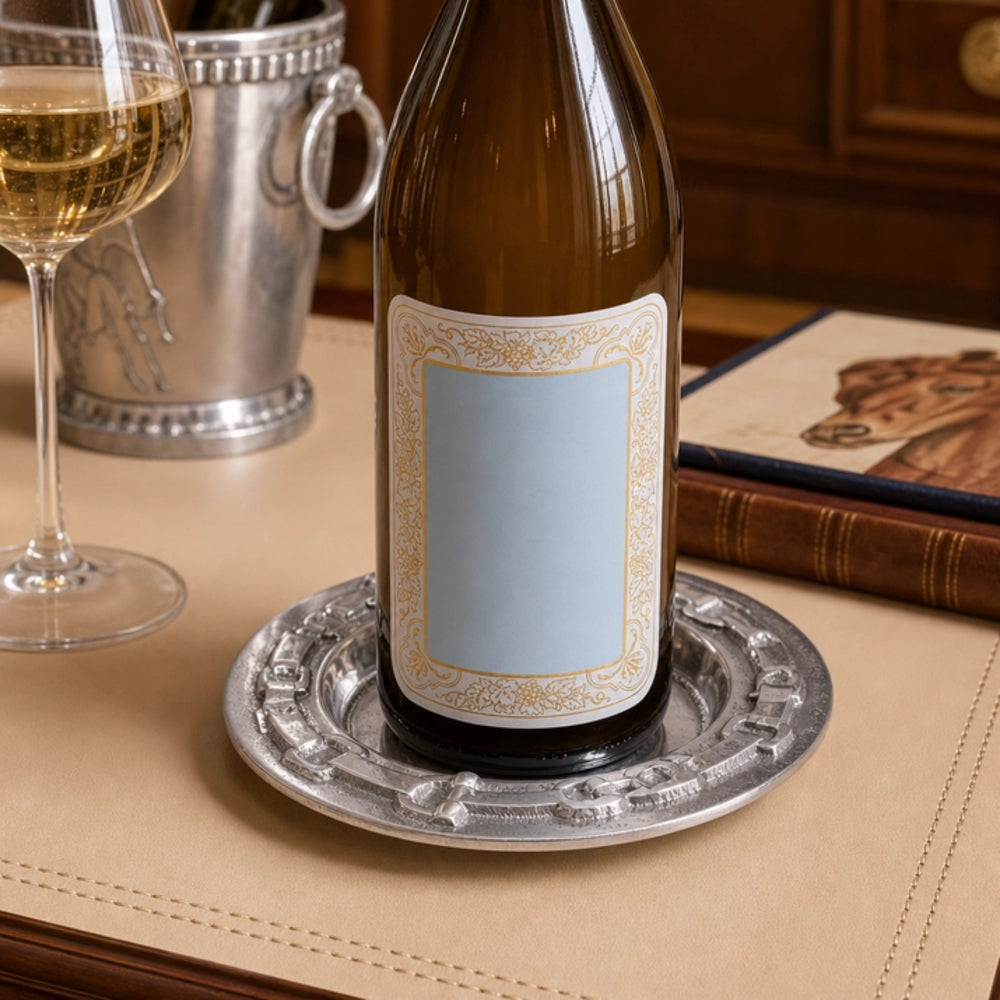 An empty brown glass wine bottle with a blank light blue label sits on an ornate silver charger plate on a beige table. A glass of white wine, a silver bucket, and a book are blurred in the background.