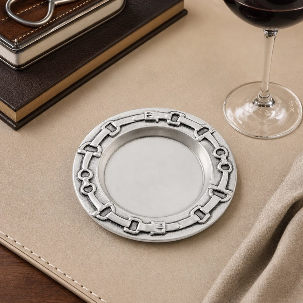 A round silver-toned metal tray with an embossed chain-link border, displayed on a beige leather surface next to a wine glass, books, and a napkin.