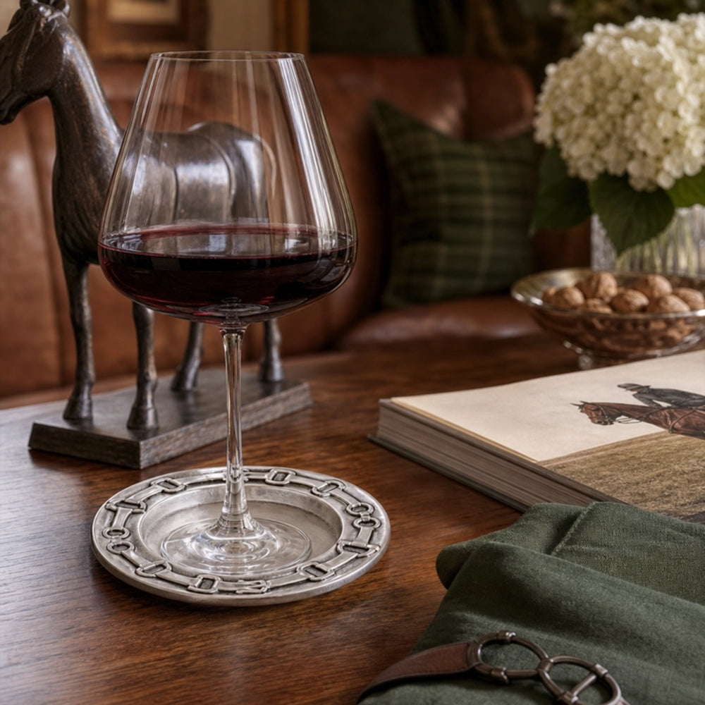 A clear stemmed wine glass partially filled with red wine sits on a round silver coaster on a wooden table in a cozy indoor setting with a leather sofa, horse figurine, and flowers in the background.