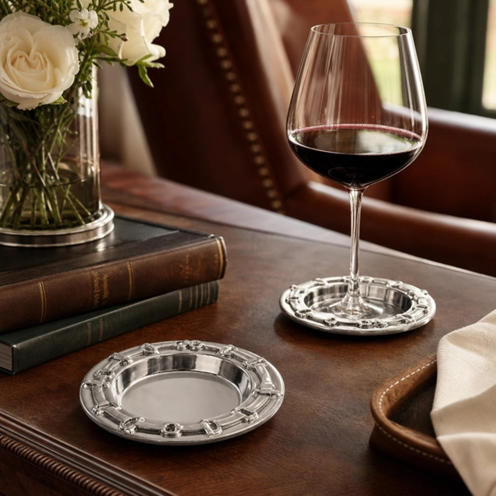 Two ornate silver coasters on a dark wooden table, with one holding a glass of red wine and the other empty, styled with books, a vase of white roses, and a leather chair in the background.