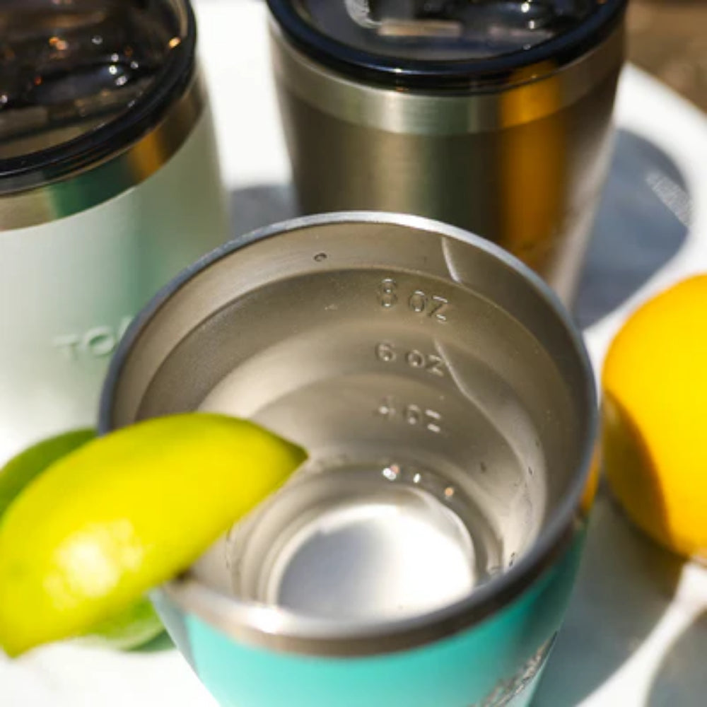 Close-up of a teal tumber with a lemon wedge, surrounded by other thermoses and lemons on a white surface.