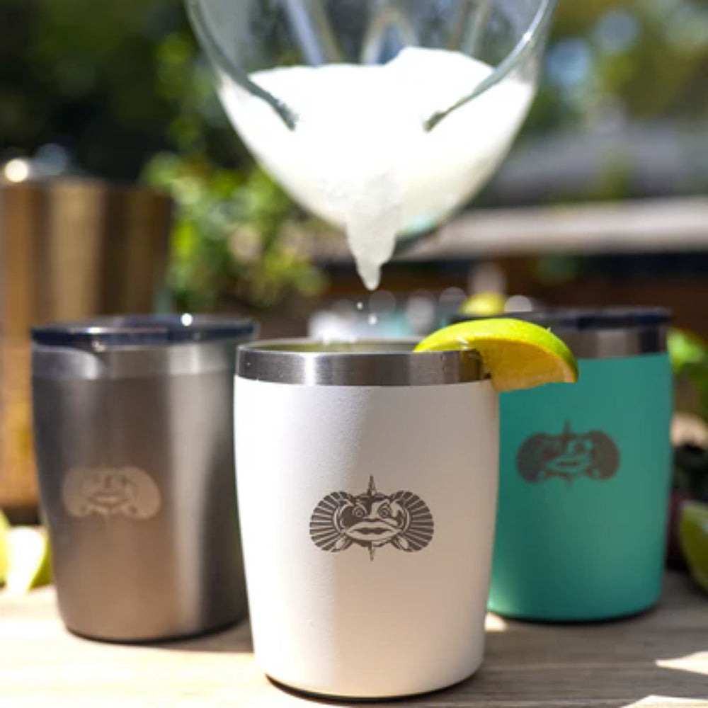 White, graphite & teal  insulated tumblers with a Toadfish logo, surrounded by other insulated cups and a glass with ice and lemon, on a wooden surface outdoors.