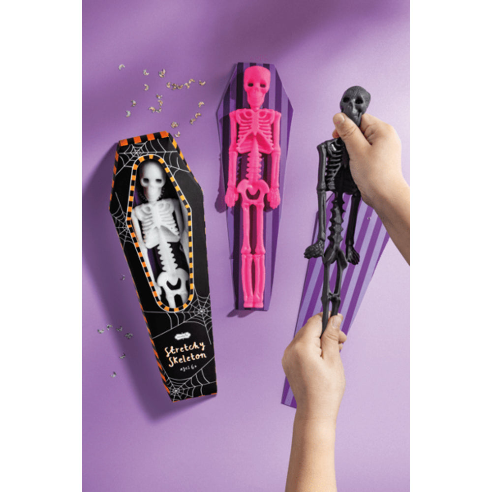 Halloween-themed skeleton toys including a coffin, pink skeleton, and black skeleton held by hands against a purple background.