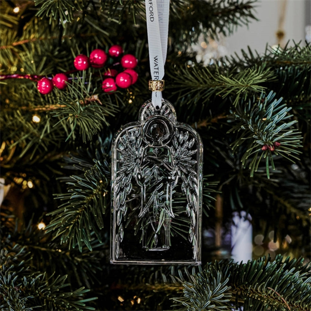 A silver-toned metal Christmas ornament shaped like an angel with wings, hanging from a white ribbon printed with the word 'WINTER' and gold lettering, displayed on a decorated Christmas tree with green branches and red berries.