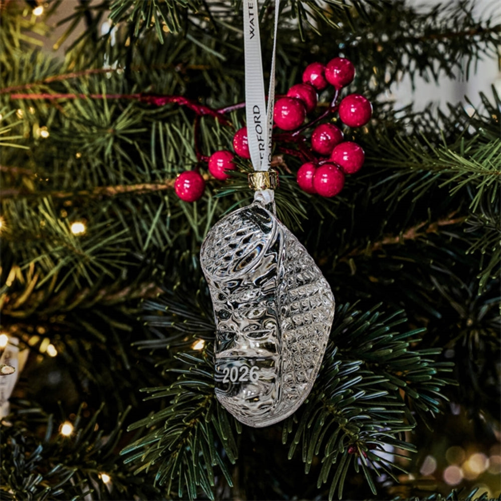 A clear glass ornament shaped like a sneaker, hanging from a white ribbon printed with the name 'WATford', displayed on a decorated Christmas tree with green pine needles and red berries.