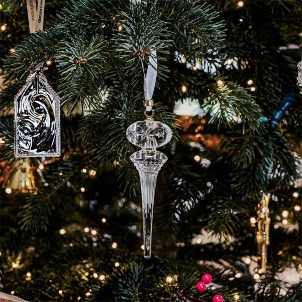 A clear faceted crystal Christmas ornament shaped like a candlestick or teardrop hangs from a green evergreen tree with warm string lights and other ornaments in the background.