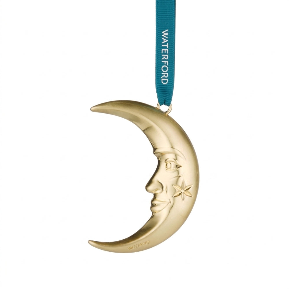 A gold crescent moon ornament with a celestial face and star detail, hanging from a teal ribbon printed with the brand name 'WATERFORD'.