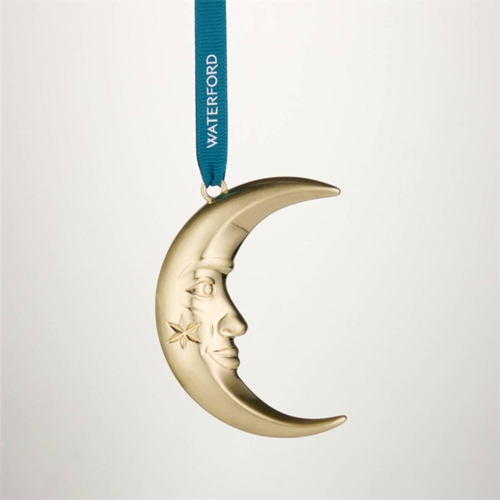 A gold crescent moon ornament with a carved face and star detail, hanging from a blue ribbon printed with the brand name WATERFORD, set against a light gray background.