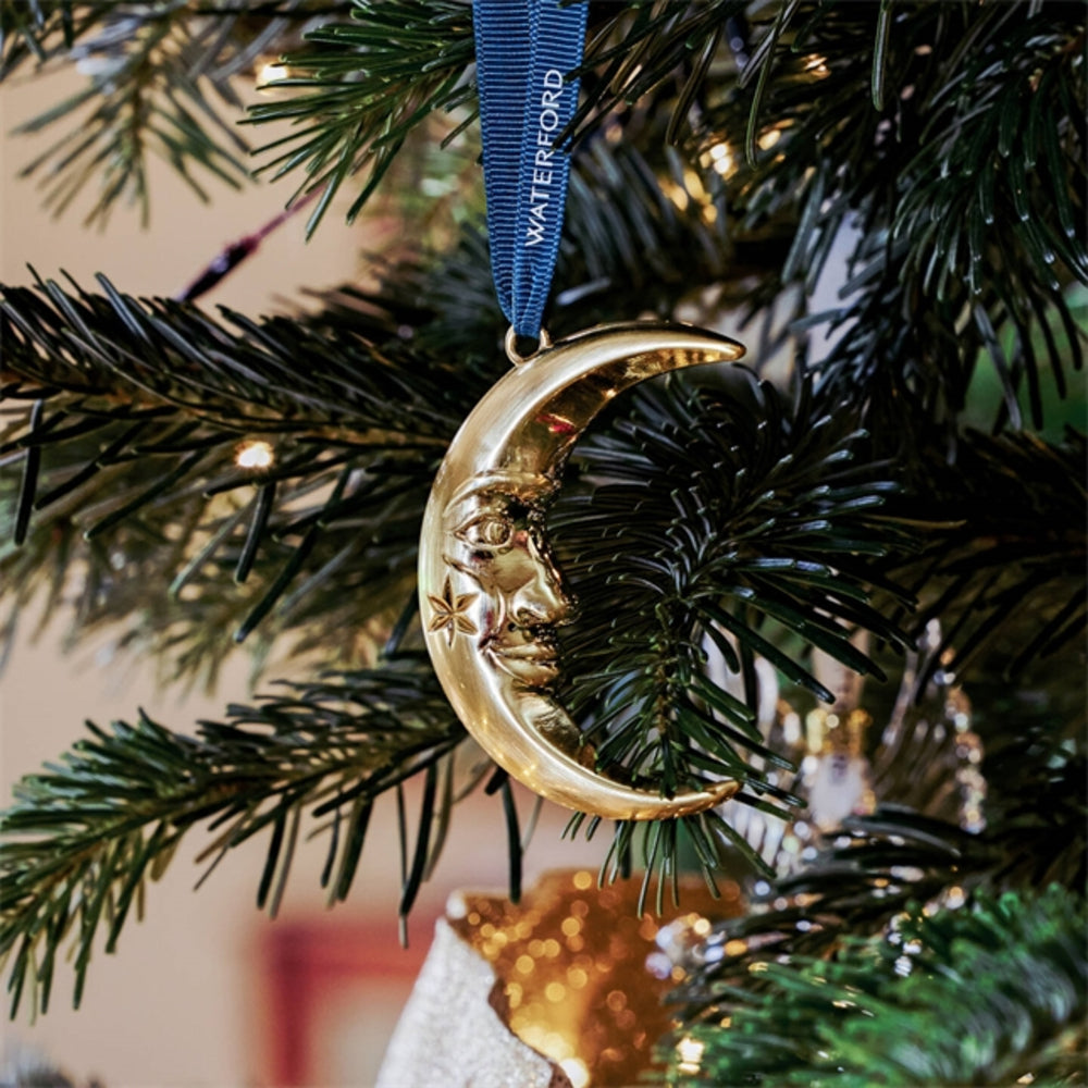 A gold crescent moon Christmas ornament with a face and star detail, hanging from a blue ribbon printed with the name 'WATERFORD', nestled among dark green pine tree branches with warm string lights.