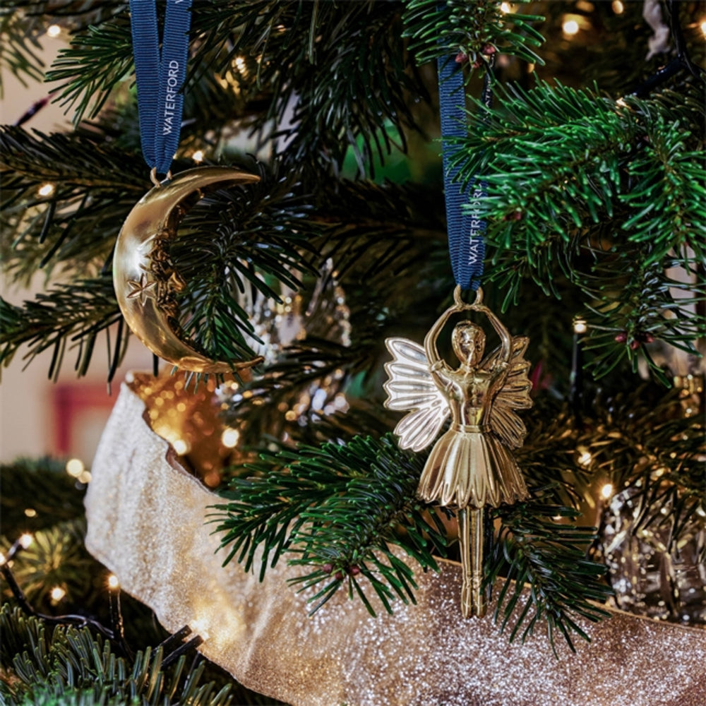 A gold angel and crescent moon Christmas ornament hanging on a decorated Christmas tree with green branches, warm string lights, and a glittery gold stocking.