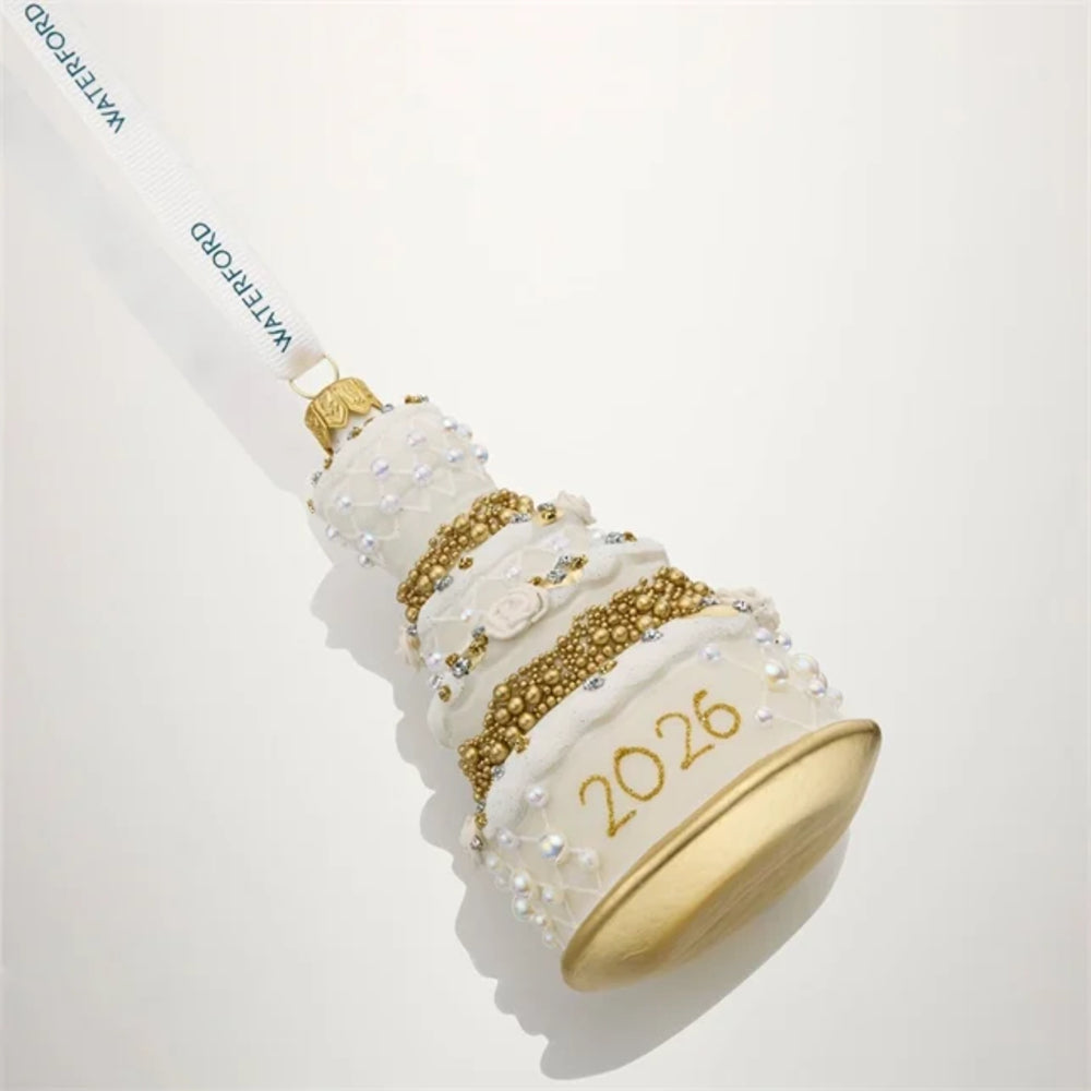 A white and gold tiered cake-shaped Christmas ornament with '2026' written in gold on the base, decorated with white pearls and gold bead details, hanging from a white ribbon printed with 'WATERFORD'.