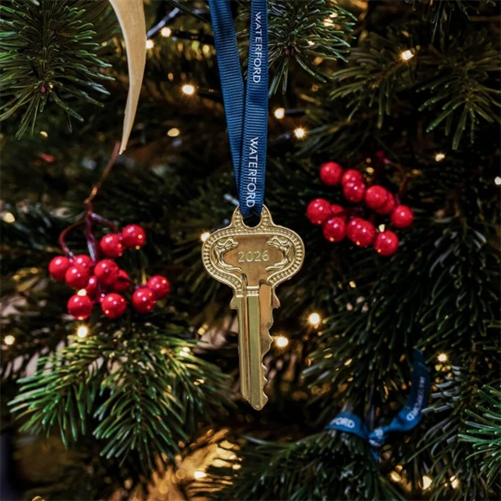 A decorative key-shaped Christmas ornament hanging from a blue ribbon printed with the name 'WATERFORD' and the year '2026', displayed on a lit Christmas tree with red berries.