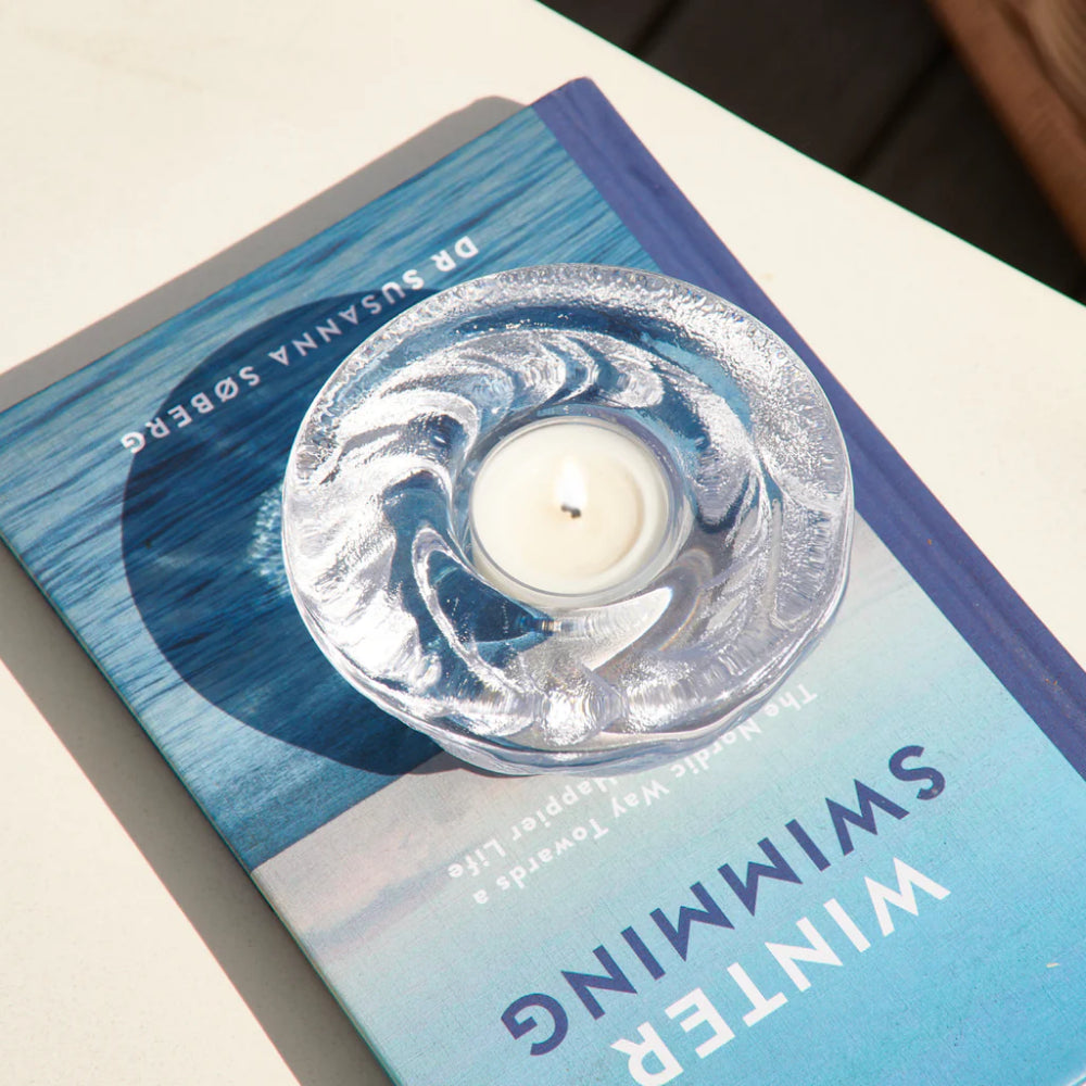 Candle in a glass holder on top of a blue book titled 'Swimming in Winter' by Susanna Sogberg.
