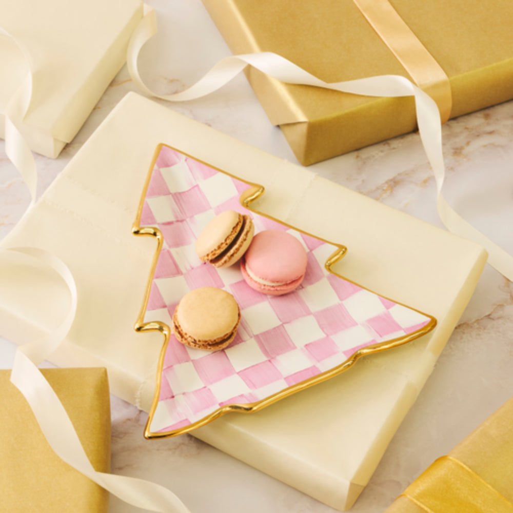 A Christmas tree-shaped ceramic plate with a pink and white checkered pattern and gold trim, styled on top of cream gift boxes with gold wrapping paper and white ribbons. Three small macarons rest on the plate.