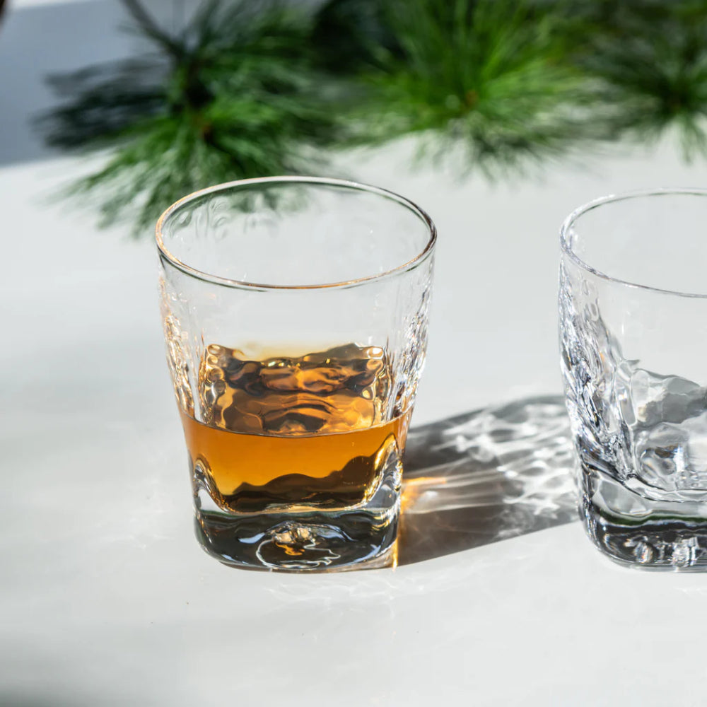 A set of two glass shot glasses on a white backround with a evergreen branch and one glass with bourbon in it.