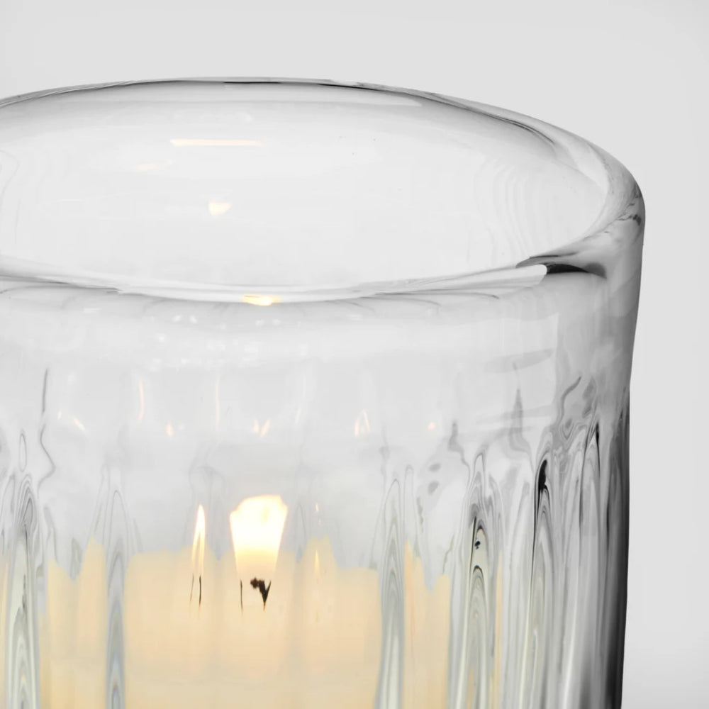 Close-up of a clear ribbed glass candle holder with a lit cream-colored candle inside, set against a plain light gray background.