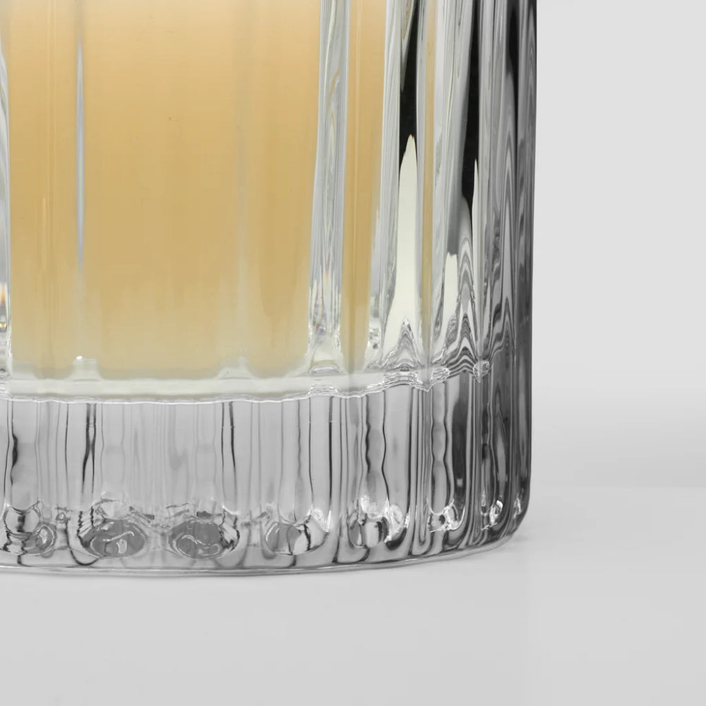 Close-up of a clear ribbed glass tumbler with a pale amber drink inside, photographed on a clean light gray background.