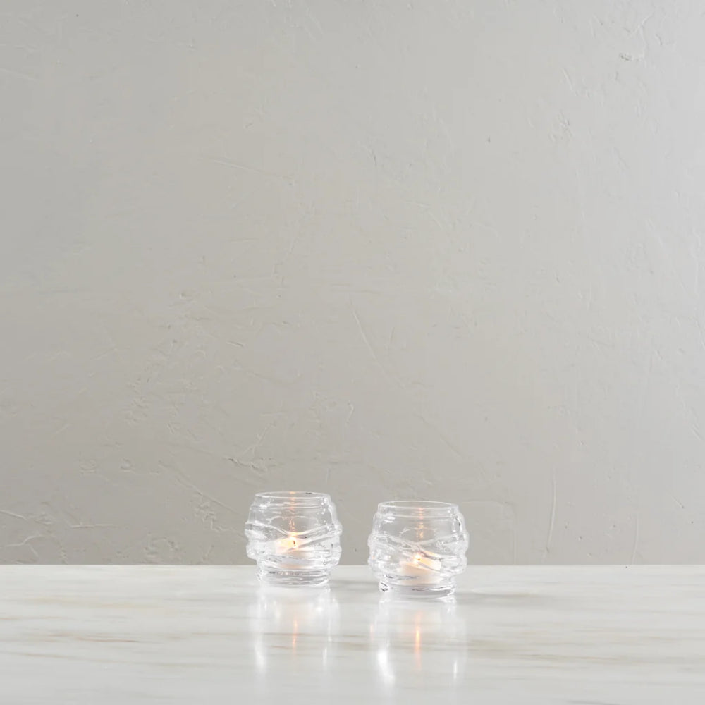 Two small clear glass candle holders with a textured, wavy pattern, each containing a lit tea light candle, set on a light marble surface against a neutral textured gray wall.
