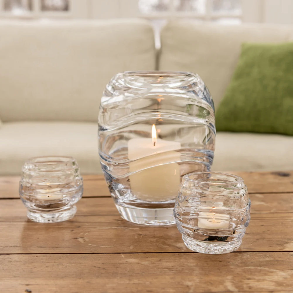 Set of three glass candle holders with candles on a wooden surface, blurred sofa in the background